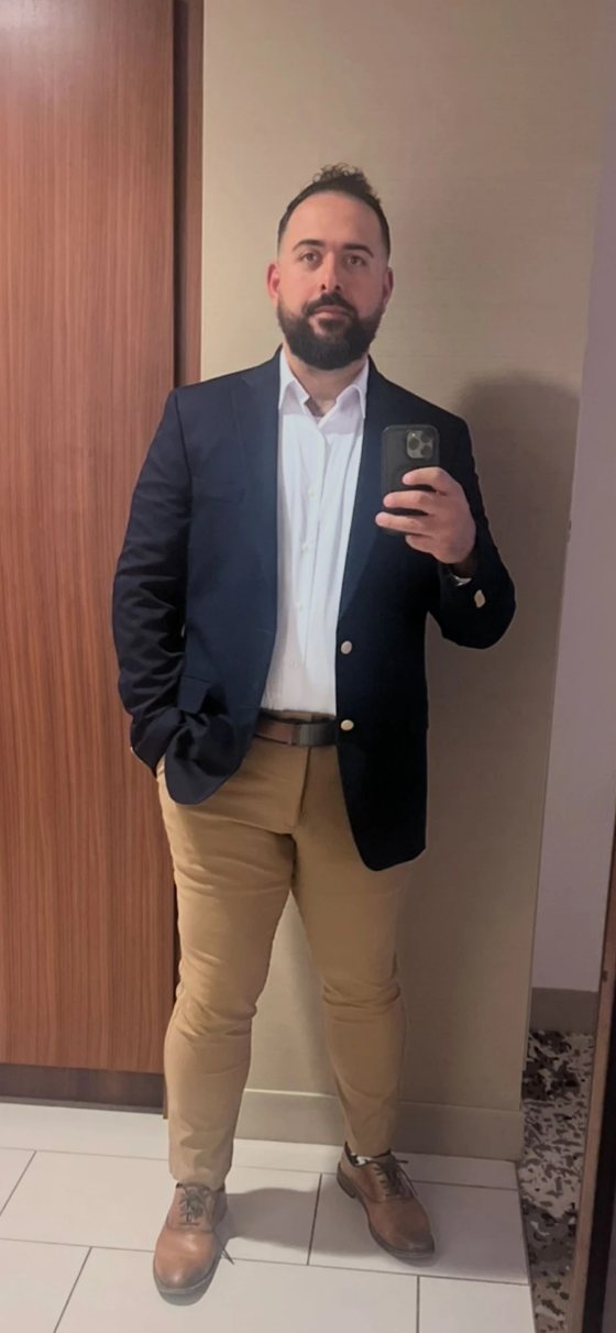 David Bechara in a navy blazer, white shirt and khaki trousers, taking a mirror selfie.