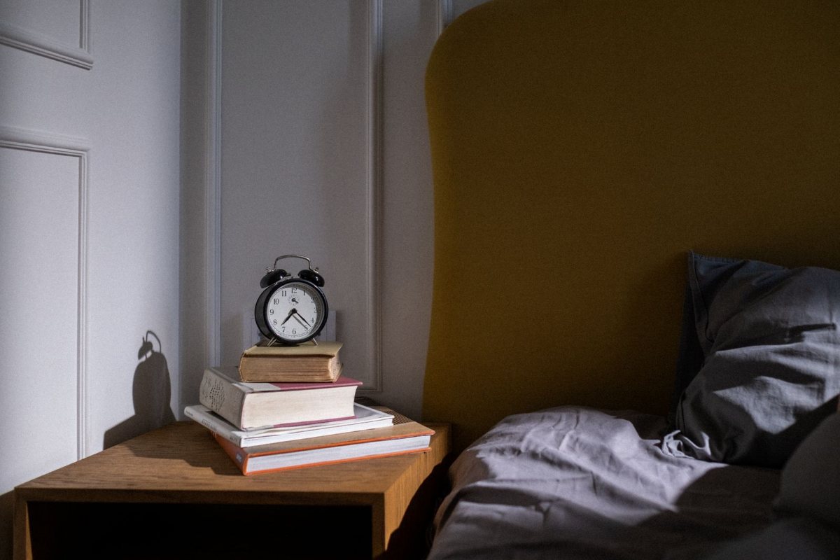 An alarm clock on a stack of books on a nightstand beside a bed.